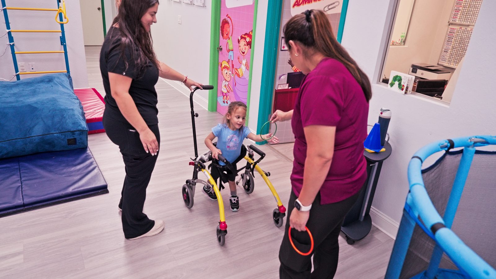 Two staff members supporting a young girl walking with a gait trainer in the therapy gym at Peek A Boo Pediatric Care in Hollywood, FL