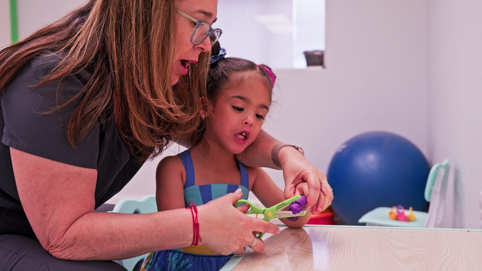 Therapist guiding a young girl's hands while she uses child-safe scissors at a table at Peek A Boo Pediatric Care in Hollywood, FL