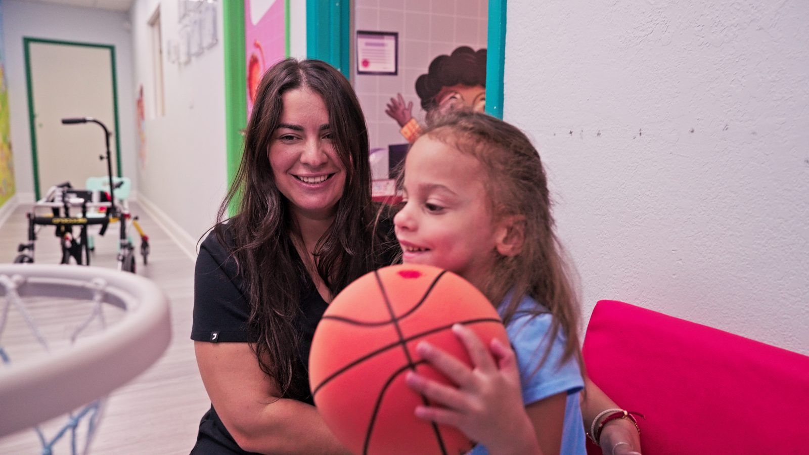 Smiling therapist and young girl holding a basketball together during a therapy session at Peek A Boo Pediatric Care in Hollywood, FL