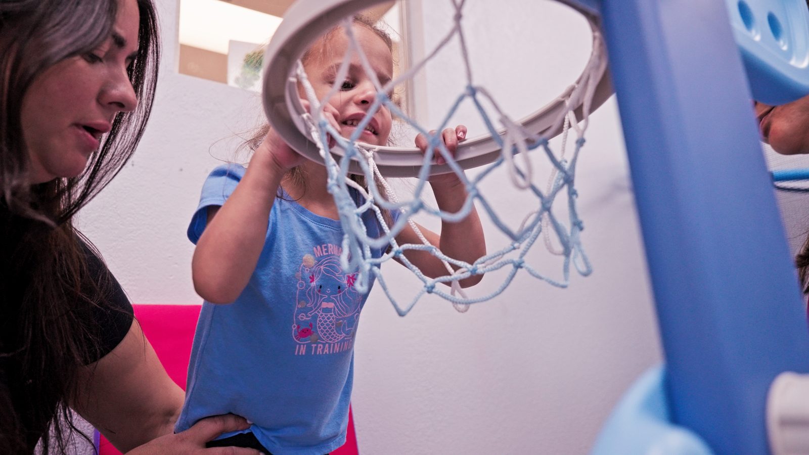 Therapist supporting a young girl as she reaches up to a basketball hoop during a therapy session at Peek A Boo Pediatric Care in Hollywood, FL