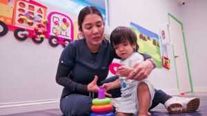 Therapist sitting on a mat with a toddler playing with a ring stacker at Peek A Boo Pediatric Care in Hollywood, FL